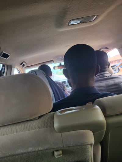 The image captures the interior of a car, likely a minivan or SUV, from the perspective of a rear passenger. The scene appears to be in Nyanya, Nigeria. In the foreground, the headrests of the car's seats are visible, upholstered in beige fabric. Behind them, three individuals are seated in the back. The person in the immediate foreground is seen from the back of their head, with short, dark hair. They are wearing a dark blue shirt. Next to them, to their right, is another person whose head and shoulders are visible. This individual has dark skin and is wearing a plaid shirt. Further to the right, the back of a third person's head is visible, with short, dark hair. This person is wearing a light-colored, checkered shirt. Through the car windows, a blurry outdoor scene is visible, suggesting the car is in motion. The car's interior features a beige headliner, a rearview mirror, and a light fixture on the ceiling. The overall impression is of a group of people traveling together in a vehicle, likely on a road in Nyanya.