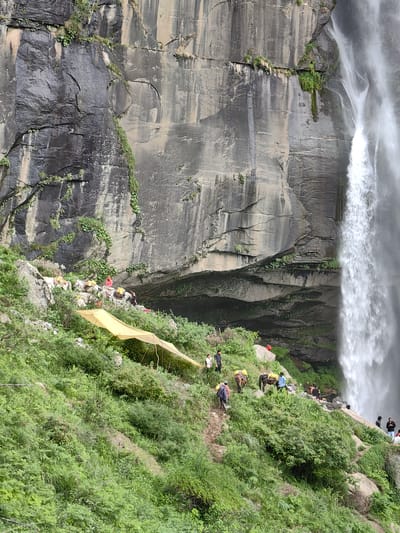 The image depicts a scene at the scenic waterfall in Bahang, India, nestled against a towering, sheer rock face. A powerful waterfall cascades down the right side of the frame, its white spray contrasting with the dark, textured rock. The rock face is mottled with shades of grey and brown, with patches of green vegetation clinging to its surface. In the foreground, a steep, verdant slope is covered in lush green foliage, with a winding dirt path visible.
Several people and pack animals are present on the slope. In the middle ground, a group of donkeys or mules, laden with yellow saddlebags, are being led by individuals in casual clothing. Some of these animals are positioned along the path, while others are further up the slope. A bright yellow tarp, supported by poles, is set up on the hillside, possibly as a shelter or for vendors.
Further down the slope and closer to the base of the waterfall, a few more individuals are visible, some standing and others seemingly engaged in activities near the water's edge. The environment is natural and rugged, with the emphasis on the grandeur of the waterfall and the surrounding mountainous terrain.