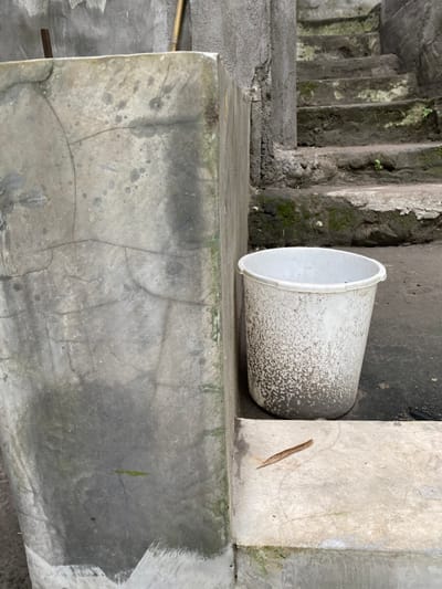 The image shows a white plastic bucket sitting on a concrete surface. The bucket is positioned between two concrete walls. The bucket has a weathered, dirty appearance. The surface the bucket sits on appears to be part of a set of concrete steps that extend upward and out of the frame. There is some green moss growing on the steps. The scene appears to be in an outdoor location. There are no people in the scene. The location is in Kecamatan Piyungan, Indonesia, at latitude -7.825 and longitude 110.453.