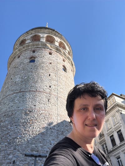 The image is a self-portrait taken outdoors in daylight, likely in the Beyoğlu district of Istanbul, Turkey. In the foreground, a person with short, dark hair and a black t-shirt is smiling at the camera. A pair of glasses rests on their t-shirt. Behind the person and to the left, the Galata Tower, a medieval stone tower, dominates the frame. The tower is made of light-colored stone with arched openings near the top and is illuminated by sunlight. To the right of the tower, a multi-story building with ornate white architectural details and windows can be seen. The sky is clear and blue.