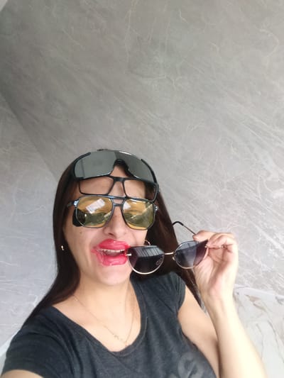 A woman is posing for a selfie-style photograph, wearing multiple pairs of sunglasses and what appears to be a very thick, bright red lipstick. She has two pairs of sunglasses perched on her face and another pair held in her mouth and right hand. She is wearing a dark gray t-shirt and a gold necklace. The background is a light gray wall with marble-like patterns. The location is unknown. The presence of Andreyevo, Russia is unverifiable.