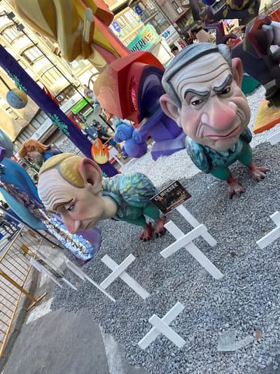 This image captures a scene in Alicante, Spain, featuring large, colorful, caricature-like figures that appear to be part of a street festival or display, possibly a "Falla" celebration, given the context of Alicante. The figures are exaggerated and whimsical, with one depicting an older man with grey hair and a stern expression, and another a creature-like figure with large ears and a smiling, slightly mischievous face. They are made of vibrant materials, with detailed sculpting and painting, suggesting they are handcrafted works of art.
The figures are positioned outdoors, set against a backdrop that includes buildings with visible storefronts and signage, indicating an urban setting. The ground is covered in gravel and asphalt, with several white cross-like structures placed around the figures, possibly as part of the display or for crowd control. In the background, glimpses of people can be seen, suggesting the presence of an audience or participants in the event.
A notable detail is a small, black sign held by one of the figures, which contains text in what appears to be Spanish. The text is difficult to read clearly in the main image but appears to be "29 MARZO INVITAN A LA MESA DE ESTOS DOS". This suggests a specific date, "March 29th," and an invitation to "the table of these two," possibly referencing the figures or a related event. The overall atmosphere seems festive and lively, typical of a Spanish cultural celebration. The lighting suggests it might be daytime, with overcast conditions or shadows from surrounding structures.
