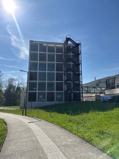 The image depicts a daytime outdoor scene on the Azurém Campus of the University of Minho in Guimarães, Portugal. A multi-story, six-level building with a light-colored concrete grid facade and alternating dark reddish-brown panels and reflective windows occupies the central and right-midground. An external black metal fire escape staircase with multiple landings is affixed to the right side of this building. To the left of the main building, a dark, contemporary streetlight stands. In the foreground, a curved concrete pathway with a central tactile paving strip composed of parallel longitudinal ridges extends from the lower left towards the upper center. A vibrant green, somewhat overgrown grassy area with scattered small purple flowers separates the pathway from the building. In the background, to the right of the main building, another modern architectural complex is visible, connected by an elevated pedestrian bridge. Several white commercial vans and other vehicles are parked near these background structures. The sky is clear blue with a prominent sunburst and lens flare originating from the upper left, indicating bright daylight. A faint white contrail from an aircraft is visible in the upper right quadrant of the sky. No individuals are prominently visible in the immediate foreground.