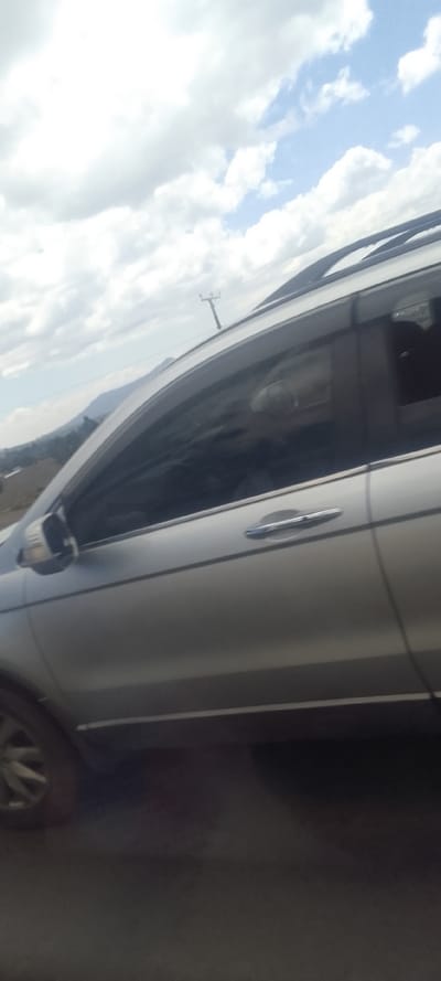 The image depicts the left profile of a silver sport utility vehicle (SUV) in Hells Gate ward, Kenya, presented with a slight tilt. The SUV features roof rails, dark rain deflectors on all side windows, and chrome door handles. Through the tinted rear passenger window, a faint, indistinct reflection of what appears to be a human head or face is discernible. The background shows a partly cloudy sky with prominent white and grey clouds against patches of blue. Distant, undulating hills are visible beneath the skyline. A wooden utility pole with a crossarm stands in the mid-ground behind the vehicle. The immediate foreground is a dark, blurred surface consistent with a road.
