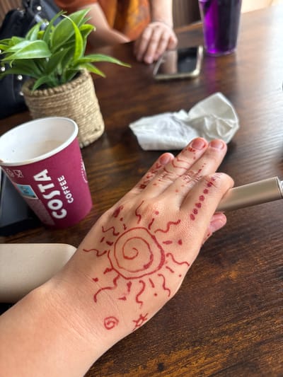 A close-up shot shows a person's hand with henna art on a wooden table. The henna design includes a sun with radiating lines and dots along the fingers. A Costa Coffee cup is visible to the left of the hand, and a metallic object, possibly a vape pen, is being held by the hand. In the background, a small potted plant, a phone, and a glass of purple liquid are present. The overall setting appears to be a casual indoor environment, likely a cafe or a home. There is no information in the image to suggest the location is Garmen, Bulgaria.