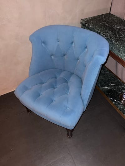 A light blue upholstered armchair with tufted detailing is positioned in the foreground, angled towards the viewer. The armchair has a curved back and a rounded seat. It rests on four dark, short legs. To the right of the armchair, a two-tiered marble table with a dark green, veined surface is visible. The table has thin wooden trim around its edges and appears to be part of a larger furniture set. The background consists of a textured, pale beige wall. The floor is a dark, matte surface, possibly concrete or dark tile. The scene is lit from above, casting soft shadows. No people are present. The location is an indoor setting, possibly a room in a residential or commercial establishment in Vyshen'ky, Ukraine.