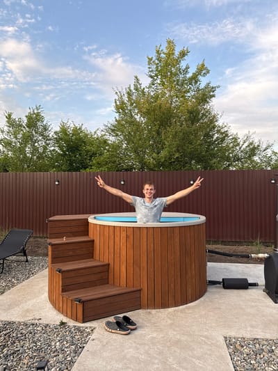 The image shows a man standing in a wooden hot tub with blue water. He is smiling and holding up two fingers in a peace sign. He is wearing a grey t-shirt. The hot tub is surrounded by wooden decking and steps. To the left of the hot tub is a brown fence and a large green tree. To the right of the hot tub is a concrete patio with gravel and a black lounge chair. There are a pair of black crocs with red stripes on the patio. The sky is blue with some white clouds. The time of day appears to be late afternoon or early evening. The weather is clear and sunny. The location is Starokorsunskaya, Russia.