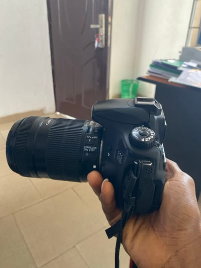 A dark-skinned right hand, with a visible white fingernail on the index finger, grips a black Canon EOS 60D DSLR camera. An attached black Canon EF-S 18-135mm lens, set to 'AF' (Autofocus) and 'STABILIZER ON,' extends forward. The camera's mode dial is visible, positioned towards the top. The scene is an indoor environment within a building in Akarakumoh, Nigeria. The flooring consists of light-colored, square tiles. In the background, a closed brown wooden door with a silver handle and a key attached is prominent on the left. To the right of the door, a white wall is visible. Further right, a dark desk with stacks of papers and a partially obscured green waste bin are present.