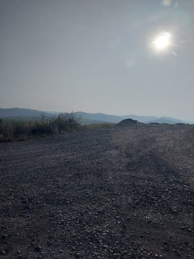 The image depicts a landscape with a bright sun in the upper left corner, casting lens flares and illuminating a hazy sky. The foreground is dominated by a dark, gravelly surface, appearing to be a road or pathway. To the left of this gravel path, a strip of vegetation, including grasses and shrubs, is visible. Beyond the vegetation, a series of rolling hills fades into the distance, layered with a soft, blue haze. The scene suggests an outdoor setting during the daytime, possibly late morning or early afternoon given the sun's position and intensity. There are no visible people or text in the image. The overall mood is one of a serene, rural landscape. The orientation of the image is rotated 90 degrees counter-clockwise.
