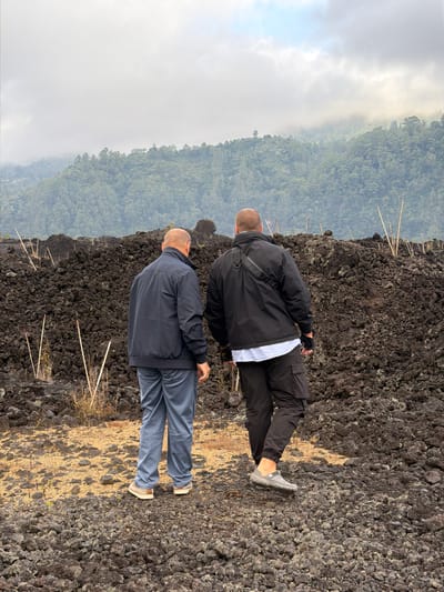 Two bald men are standing in a field of volcanic rock in Kintamani, Indonesia. The man on the left is wearing a dark blue jacket and grey pants. The man on the right is wearing a black jacket, black pants, and grey shoes. In the background, there is a lush green mountain covered in trees, with a cloudy sky above. The volcanic rock field is a mix of dark grey and black rocks, with some dry grass and thin wooden sticks scattered throughout. The scene appears to be a natural landscape, possibly a hiking trail or an area affected by a volcanic eruption.