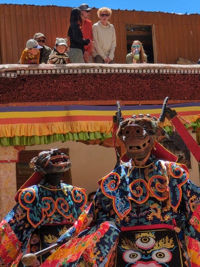 In Pibiting, India, two individuals in elaborate, traditional costumes and masks are the central focus. The person on the right wears a dark, horned mask with a snarling expression and a richly colored, blue robe adorned with golden dragons and swirling patterns. The person on the left wears a black mask with an open, jagged mouth and a similar colorful robe. Both figures appear to be performing or participating in a ritual, holding staffs.
In the background, a multi-tiered structure is visible, with colorful fabric draped across it in horizontal bands of yellow, orange, red, and blue. Several spectators, including children and adults, are observing the event from a balcony-like area above the performers. The architecture suggests a religious or cultural festival setting. The sky above is clear and blue.