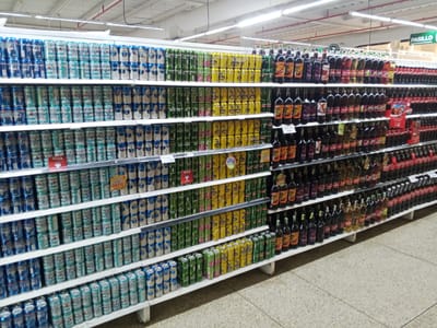 The image shows a supermarket aisle in El Valle del Espíritu Santo, Venezuela, filled with shelves of beverages. The aisle is brightly lit by fluorescent lights. The shelves are stocked with numerous cans and bottles of beer, soda, and other drinks. On the top shelf, there are rows of beer cans, predominantly in blue and silver. Below this, more beer cans continue, in various colors including blue, green, and yellow. Further down the shelves, bottles of wine and other alcoholic beverages are displayed. Some of these bottles are dark brown, while others are clear with colored liquids. Towards the right side of the image, rows of red Coca-Cola bottles are visible. The floor is a tiled surface. There are no people visible in the image. The shelves are organized and neatly stacked with products, indicating a well-maintained retail environment. A green sign with white text that reads "PASILLO" is visible hanging above the shelves on the right side of the image.