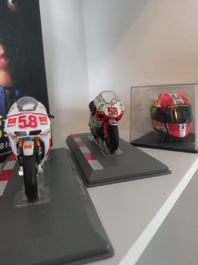 The image shows a display of motorcycle memorabilia, likely miniature models, arranged on a white shelf or wall. The items include two miniature motorcycle models mounted on gray stands with red and white striped sections, and a helmet in a clear display case. The number "58" is visible on both motorcycles. The photograph appears to be taken at an angle, giving a slightly tilted perspective of the display. Text is visible on the stands and possibly on the motorcycle models, including brands like "DELLORTO", "MIDAC", "Getis", and the motorcycle model "HONDA RC212V (2011)". The location in the city of Crauthem, Luxembourg is not discernable from the image alone.
