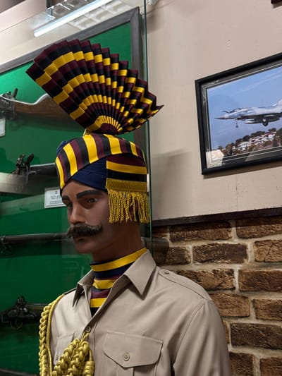 A mannequin stands inside a glass display case. It wears a khaki uniform and a turban with yellow, burgundy, and navy blue stripes. A pleated fan, mirroring the turban's colors, is positioned beside the mannequin's head. Antique rifles are visible within the display. A framed photograph of an aircraft hangs on the wall above, with a brick wall on the right.