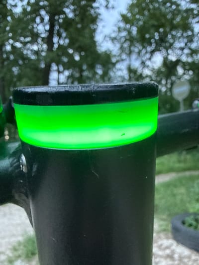 The image shows a close-up of a black bicycle frame with a vibrant green accent. The green component appears to be a decorative band or a reflective strip, possibly for increased visibility. The texture of the black frame suggests it is made of metal, with some subtle scuff marks visible. The background is blurred and out of focus, but it hints at an outdoor setting with greenery and possibly a path or gravel.
The composition is a tight shot, focusing entirely on the bicycle's frame and the striking green detail. The lighting suggests it's daytime, but the exact time is unclear due to the lack of strong shadows. The overall impression is that of a detail shot highlighting a specific feature of the bicycle. There is no text visible in the image. The context provided indicates the location is Irpin, Ukraine.