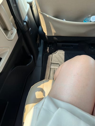 The scene depicts the interior of a passenger vehicle, located in the city of Veracruz, Mexico. A human thigh and knee, exhibiting light-toned skin, occupy the lower central and right portion of the frame, resting on a tan-colored seat. The limb is partially covered by a light-colored garment. To the left, a two-toned door panel, featuring a light upper section and a dark lower section, includes a recessed storage compartment. The back of a front seat, upholstered in tan material, is visible in the upper right; its rear storage pocket contains a clear plastic water bottle with a white cap and other small, indistinct items. The vehicle's floor, covered by a dark rubber floor mat with a geometric pattern, is visible beneath the front seat. Strong direct sunlight illuminates parts of the interior, casting defined shadows on the leg, seat, and floor.