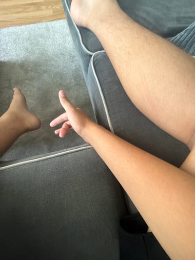 A top-down view shows a partially visible human body on and around a gray upholstered sofa, with a light gray rug partially visible. A lower leg and foot are situated on the rug next to the sofa. An arm extends from the sofa, with a hand hovering above the rug. The left leg, also partially visible, rests on the sofa cushion. The scene includes a glimpse of light-colored wooden flooring in the lower left corner. Given the prompt, the implied location is Gorni Bogrov, Bulgaria.