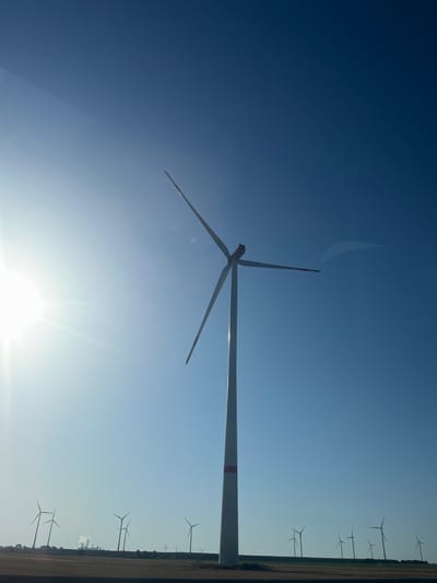 The image shows a wind farm in the city of Jüchen, Germany, under a clear blue sky. A large, prominent wind turbine stands in the foreground, its blades angled upwards. In the background, a row of smaller wind turbines are silhouetted against the horizon, along with distant industrial structures emitting smoke. The sun is shining brightly on the left side of the frame, casting a lens flare. The ground appears to be a field of dry vegetation. There are no people visible in the image.