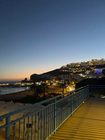 The image shows a cityscape at dusk in Puerto Rico, Spain. The sky is a gradient of dark blue to orange, with a faint crescent moon visible. The city is built on a hillside, with numerous buildings lit up in the night. A metal railing and wooden walkway are in the foreground, suggesting a viewpoint or elevated area. Palm trees and other vegetation are interspersed among the buildings. Below, there is a beach and harbor area with boats and artificial beach, illuminated by streetlights.