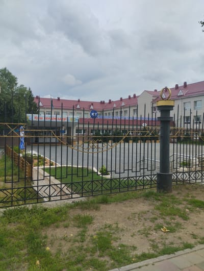 The image shows the exterior of a building with a red-tiled roof and beige walls. In front of the building is a black metal fence with decorative gold elements. A sign above the entrance reads "КІШ КЕЛДІҢДЕР!". The building appears to be a public institution, possibly a school or administrative building, located in Kostanay, Kazakhstan. The sky is overcast. There is a paved area behind the fence, leading to the entrance of the building. Green grass and some trees are visible on the left side of the image. A lamppost with a golden ring on top stands to the right of the fence.