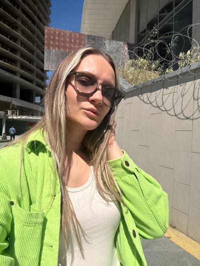 A woman wearing sunglasses, a white top, and a bright green corduroy jacket poses for a selfie in Esenyurt, Turkey. In the background, there's a building under construction and a concrete wall topped with razor wire. The woman is facing the camera, partially angled, with sunlight casting shadows on her face and clothing.
