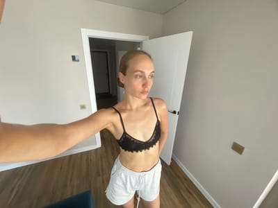 A young woman with blonde hair pulled back stands in a brightly lit room, taking a selfie with her left arm extended. She is wearing a black lace bralette and light grey sweat shorts with an orange logo on the right leg. The room has light beige walls and a wooden floor. To her left, a doorway leads to another room with a darker interior and a closed door. To her right, a white door is ajar. A light switch or outlet is visible on the wall behind her, and another rectangular object is mounted on the wall to the right. The overall scene suggests a casual, domestic environment. The location is identified as Podshivalovo, Russia.