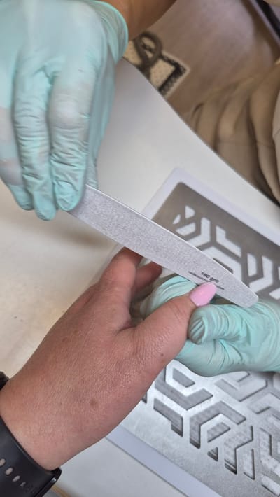 A close-up, overhead shot shows a person wearing light blue nitrile gloves filing a light pink acrylic fingernail with a grey emery board. The emery board is labeled "180 grit". The hand being worked on is also wearing a light blue nitrile glove on the thumb. The background is a white surface with a silver geometric stencil pattern. The image does not provide any information about the city of Daugavpils, Latvia, or its location within it.