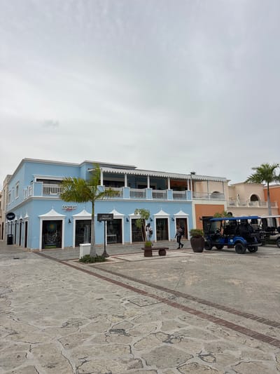 The image depicts a street scene in Higüey, Dominican Republic, likely within a commercial or resort area. A light blue, two-story building with white trim and balconies is prominent in the foreground, housing several shops with awnings. A single palm tree stands in front of the building, and a "CALLE COMERCIAL" (Commercial Street) sign is visible. To the right, other buildings in shades of peach and cream are partially visible, featuring similar architectural elements. Several golf carts, one prominently blue and the others partially obscured, are parked on the stone-paved street. Two individuals are walking on the sidewalk in front of the blue building, and another is visible further down the street. The sky is overcast and light gray. The ground is paved with irregular-shaped stones bordered by a red brick pattern.