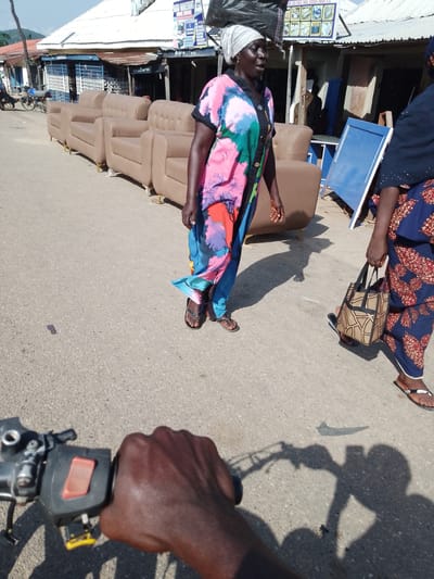 A first-person perspective shows a person holding the handlebars of what appears to be a motorcycle in Serti, Nigeria. The scene is a street lined with shops and displays of furniture, including several brown leather sofas laid out on the pavement. Two women are visible in the frame. The woman in the foreground, on the left, is wearing a vibrant, multi-colored patterned dress and a headscarf. She is walking away from the camera. To her right, another woman, dressed in a navy blue fabric with a red floral pattern, walks towards the camera, carrying a woven handbag. The ground is asphalt, and shadows suggest it is a sunny day, likely mid-morning or afternoon. The shops in the background have corrugated metal roofs and barred windows, typical of a market setting. A sign above one of the shops reads "ABU MASALAT" and lists services such as "BED SHELF TABLE KITCHEN CABINET E.T.C." The overall impression is of a bustling marketplace.