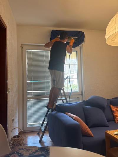 A man wearing orange gloves and a dark t-shirt stands on a ladder, cleaning an air conditioning unit above a window. The air conditioner is covered with a blue tarp. The man appears to be holding a hose or cleaning implement. The room has a blue couch with orange accent pillows, blinds on the window, and a woven basket on the floor. The scene appears to be inside a residential apartment in Sutomore, Montenegro.