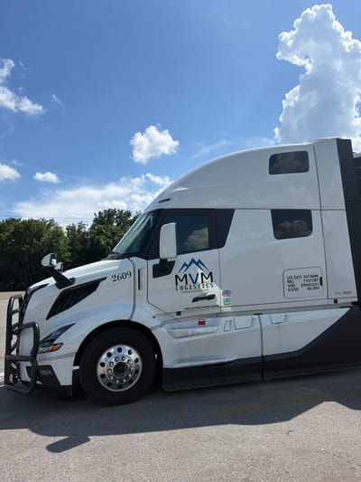 A white semi-truck is parked on asphalt under a bright blue sky with scattered white clouds. The truck has a black bull bar on the front. The side of the truck features the logo "MVM LOGISTICS VANCOUVER, WA" and various labels including "US DOT 3413699", "MC 1101387", "VIN SN693201", and "GVW 80,000". The number "2609" is also visible on the side of the cab. Trees are visible in the background. The image appears to be taken in Lebanon, United States of America.