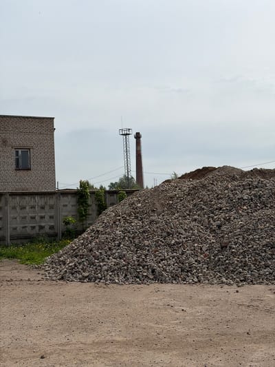 The image depicts an industrial scene in Uva, Russia. A large pile of grey gravel dominates the right side of the frame, alongside a dirt road. In the background, there's a brick building with a single window, a concrete fence with climbing vegetation, and a tall metal tower. A brick smokestack is also visible. The sky is overcast. There are no people visible in the frame.