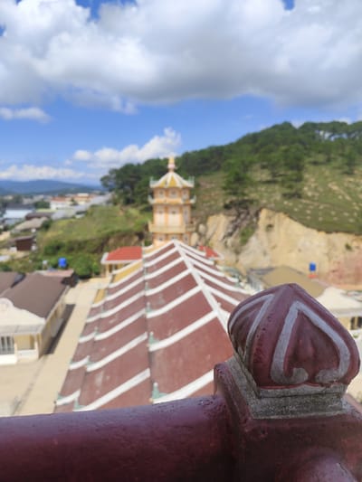 The image is a medium shot, taken from an elevated viewpoint, looking down on a temple complex and its surrounding environment. The foreground is dominated by a dark red railing and a decorative finial, which appears to be made of plaster or concrete and painted with white swirling patterns. The finial is situated on a square base.
Beyond the railing, the focal point is a large, multi-tiered pagoda-like structure with a distinctive red roof adorned with white stripes. The roof has a complex, stepped design. To the left of this structure, several buildings with cream-colored walls and brown roofs are visible.
In the mid-ground, a tall, yellow tower with multiple levels and a decorative yellow cap stands prominently. This tower also features architectural elements characteristic of East Asian temples. The buildings and the tower are situated on a hillside, which is covered with green trees and vegetation. The terrain appears to be uneven, with some exposed soil and rock faces visible.
The background reveals a hazy cityscape with more buildings and infrastructure stretching into the distance. Further beyond the city, a range of green mountains forms the horizon. The sky above is a vibrant blue, punctuated by fluffy white clouds.
The image appears to be taken in Lộc Quý, Vietnam, given the architectural style of the temple complex and the hilly, vegetated landscape often found in the region. There are no people visible in the image. The overall scene depicts a tranquil temple setting within a broader urban and natural landscape. The lighting suggests it is daytime, possibly late morning or afternoon, given the bright illumination and clear sky.