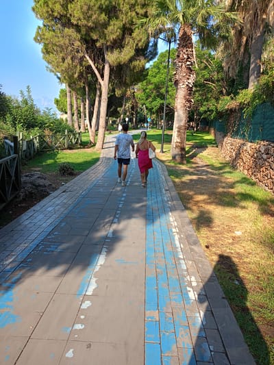 A man and a woman are walking away from the camera down a paved path in Antalya, Turkey. The man is wearing a white t-shirt with a pattern and dark shorts. The woman is wearing a bright pink dress and carrying a white bag. The path is lined with trees, including pine and palm trees, and green grass. A stone wall with a green fence runs along the right side of the path. The sun is shining, casting shadows on the path. The path is made of rectangular paving stones, with some blue paint markings. In the distance, a hazy mountain range can be seen.