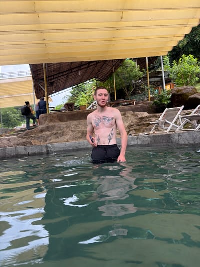 A young Caucasian male with tattoos on his chest and abdomen stands in the water of a pool, wearing black shorts. He is holding a dark-colored smartphone in his right hand. The man has short, light brown hair and a beard. In the background, two other individuals are walking up a series of stone steps. One is wearing a dark backpack, and the other is wearing a dark blue shirt. Further in the background, there is lush green foliage, two lounge chairs, and a thatched-roof structure. The scene appears to be an outdoor resort or natural hot spring area in Khanh Vinh, Vietnam. The water is clear and reflects the surrounding environment.