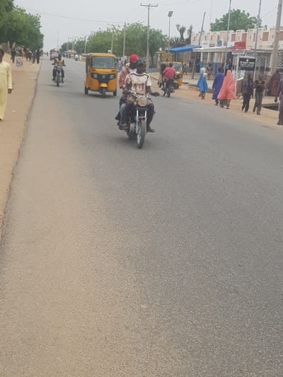 The image depicts a street scene in Dabawa, Nigeria. The road, which takes up most of the frame, is paved and appears to be made of asphalt. A motorcycle with two riders, one wearing a red hat, is traveling along the road. In the background, there's a yellow tricycle and another motorcycle, also in motion. On the left side of the road, a group of people are walking on a sidewalk in front of buildings. Some are wearing brightly colored clothing, possibly robes or wraps. Trees and utility poles are visible alongside the buildings. The sky is overcast.