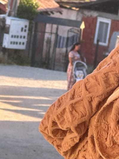 The image shows a section of tan, textured cloth in the foreground. In the blurred background, a street scene is visible. A person in a patterned top is pushing a stroller, with buildings and trees lining the street. The location is presumed to be Sandanski, Bulgaria. A building with multiple windows and a red brick building can also be seen. The image is rotated approximately 90 degrees counter-clockwise.