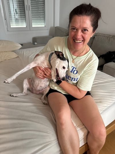 In a domestic indoor setting, a woman with brown hair pulled back in a bun, wearing a pale green t-shirt with blue text and black shorts, sits on a white-sheeted bed. She is holding a white and black-spotted dog, which is partially obscured. The woman is looking towards the camera and smiling. The dog's head is turned away from the camera, and its mouth is open. A window with blinds is visible in the background, along with a couch. The scene appears to be a bedroom. There is no specific indication of the location being the city of Murcia, Spain.