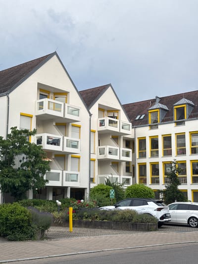 A three-story white apartment building with a brown tile roof and bright yellow window frames is visible in Sindelfingen, Germany. The building features multiple balconies with glass railings and white accents. In front of the building, a variety of green shrubs and flowering plants are present, including lavender and roses. Two white cars, an SUV and a station wagon, are parked in the paved area in front of the building. The sky is overcast and gray.