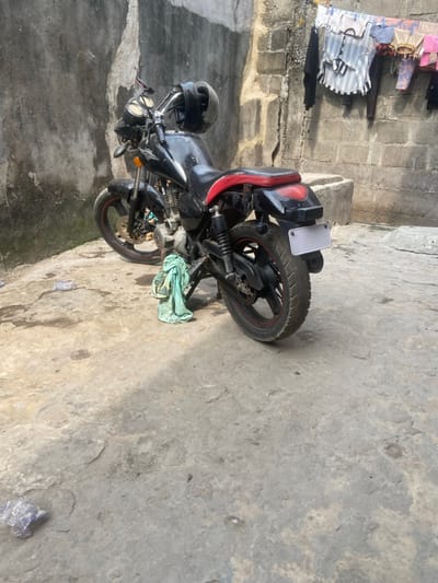 The scene depicts an outdoor concrete area within Alaiyabiagba, Nigeria. A black HOVEN brand motorcycle with a red seat and black rims with red accents is parked centrally on an uneven, stained concrete surface. A black helmet rests on its handlebars, and a light green cloth is draped over the lower engine assembly on the left side. The motorcycle's rear license plate is blank white. To the left, a weathered concrete block wall displays dark discoloration. To the right, another concrete block wall features a clothesline from which various garments, including striped shirts and other colored items, are suspended. A crumpled piece of blue material lies on the concrete ground in the lower-left foreground. No individuals are visible.