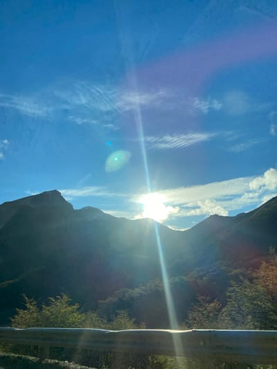 The image displays a sunlit mountainous landscape located near Lago Escondido, Argentina. A metallic road guardrail, slightly out of focus, spans the lower portion of the frame. Above the guardrail, dense green and autumnal foliage covers the lower mountain slopes. The mid-ground and background are dominated by rugged, dark mountains, their peaks appearing mostly in shadow, with subtle hints of snow or lighter rock visible on some summits. The sun, positioned behind the central peaks, emits a prominent vertical lens flare and a bright, partially obscured glow, casting strong light across the scene. The sky is a clear, vibrant blue, populated by scattered white clouds, including wispy cirrus formations and more defined cloud masses concentrated around the sun. A distinct turquoise-blue lens flare artifact is present to the left of the main sunburst. No individuals or overt actions are depicted.