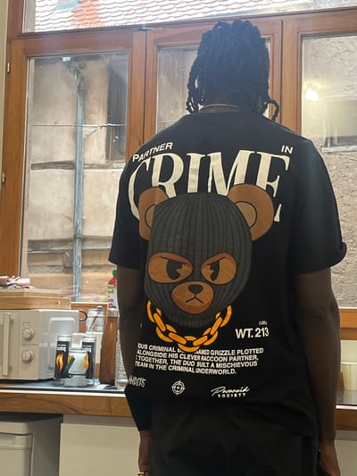 The image displays a person from behind, positioned within a kitchen interior and looking out through a multi-pane wooden window. The individual has dark, braided hair and wears a black t-shirt. A thin, gold-colored chain is visible around the person's neck. The t-shirt features a large graphic on the back with "PARTNER IN CRIME" in white capital letters. Below this text is an illustration of a brown bear-like character wearing a black knitted balaclava, revealing only its angry-looking, light brown eyes and snout. The character is depicted with a thick gold-colored chain around its neck. Text accompanying the graphic includes "WT. 213 (LBS)" and a partial narrative: "DIOUS CRIMINAL NAMED GRIZZLE PLOTTED ALONGSIDE HIS CLEVER RACCOON PARTNER, TOGETHER, THE DUO BUILT A MISCHIEVOUS TEAM IN THE CRIMINAL UNDERWORLD." Further details like "BANDITS" and "PARANOID SOCIETY" with a target symbol are also visible on the lower portion of the shirt.

On the kitchen counter to the left, a white microwave oven is present. Adjacent to it are two black cans, labeled "Burn" and featuring yellow and red chili pepper designs. Behind these, a clear glass pitcher and a brown water bottle are visible, along with a partial view of a light-colored wooden cutting board.

Through the window, the exterior environment is consistent with the historic city of Sélestat, France. Visible architecture includes a building with light-colored walls, dark timber beams, suggesting a half-timbered construction, and a traditional terracotta tiled roof in the upper left section. The view appears to be into a narrow courtyard or alleyway between old buildings.