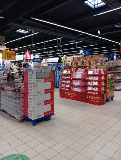 The image depicts the interior of a brightly lit supermarket, likely a Carrefour based on signage. The flooring is tiled in a light beige color, and the ceiling features a dark metal structure with numerous fluorescent lights and ventilation systems. In the foreground, a stack of boxes with red and white branding, featuring the word "VIGO!" and "LIVE GOOD" prominently, are arranged on a blue plastic pallet. To the left, several large pallets wrapped in plastic are visible, containing dark-colored products. Further back, a display of red tiered stands holds numerous smaller boxes branded with "merci". A variety of other products, including bags of snacks and bottles, are visible on shelves in the background. There are no visible people interacting with the merchandise. The overall scene suggests a typical retail environment within a commercial establishment in Wołomin, Poland.