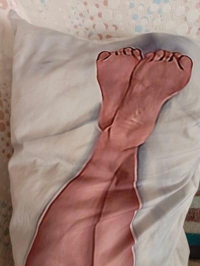 This is a close-up, high-angle shot of a pillowcase with a photorealistic print of human legs and feet. The legs are positioned in a crossed manner, with the feet prominently displayed at the top of the frame. The skin tone is rendered in shades of pink and tan, with visible toes and toenails. The fabric of the pillowcase appears to be white and slightly creased, suggesting it is a soft material. The background is partially visible and shows a textured surface with a pattern of light pink and blue circles. The scene is indoors, and the pillowcase is likely on a bed or sofa. The image provides no information about the location in Zalesovo, Russia.