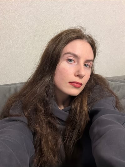 The scene, located indoors in Yanino-1, Russia, features a single adult female in a frontal, chest-up portrait. The subject has long, dark brown, slightly wavy hair that extends over her shoulders, fair skin with noticeable redness on the cheeks, light-colored eyes, and red lipstick. She is attired in a dark grey hooded sweatshirt. The background consists of a plain, light-colored, textured wall occupying the upper left and central-upper portion of the frame, and a dark grey upholstered surface, consistent with a sofa back or cushion, visible behind her shoulders and to the right. No specific actions or interactions are depicted beyond the subject's direct gaze towards the camera.