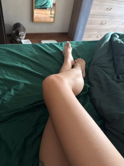 A forensic description of a bedroom interior located in Reutov, Russia, captured from an elevated perspective. The foreground features the bare lower legs and feet of an individual resting on a bed covered with dark green fabric bedding. The legs are crossed at the thighs, with visible knees and light-colored toenail polish on some toes. In the mid-background, a grey domestic short-haired cat is situated on the wooden floor, facing downward towards two small, white bowls positioned near a dark piece of furniture. A rectangular mirror with a gold-colored frame is mounted on the wall above the cat, reflecting a portion of the green bedding and a wooden surface. To the right, a light-toned wooden dresser with multiple drawers, each fitted with gold-colored pulls, is partially visible. The scene is illuminated by natural light.