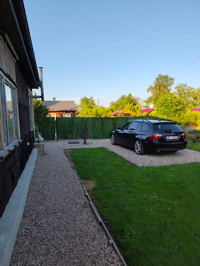 The image captures a side view of a dark-colored station wagon parked on a gravel surface next to a grassy lawn. The car, a BMW with license plate MT-9804, faces towards the right of the frame. Behind the car, a green fence runs horizontally, separating the yard from other properties. Beyond the fence, there are glimpses of wooden houses with pitched roofs and lush green trees. The foreground shows the side of a dark, weathered wooden house with visible window frames. A gravel path runs alongside the house, leading towards the parked car. The sky is clear and blue, suggesting a daytime scene. The location is identified as Jelgava, Latvia.