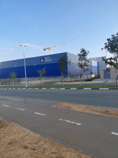 This daytime image, taken in Caesarea, Israel, captures a modern urban scene under a clear blue sky. In the foreground, a paved road with white dashed lane markings runs parallel to a dirt shoulder and a landscaped area. Along this landscaped area, several young trees are visible, some with protective stakes, and a streetlamp stands tall.
The dominant subject in the midground is a large, multi-story building featuring a striking dark blue facade. Prominently displayed on this blue section, in large white letters, is the text "& GOLF GROUP" with a stylized ampersand logo. Other parts of the building show gray paneling and reflective glass windows. The presence of two large construction cranes, visible against the clear sky in the background, indicates ongoing construction or development in the vicinity. Tree foliage also frames the upper edges of the sky. The overall scene suggests a commercial or industrial area that is actively being developed.