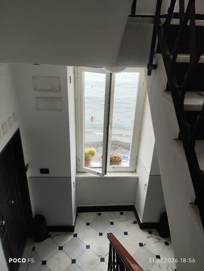 An interior shot, looking down a staircase towards a window with a view of the sea, is captured from a high angle. The staircase has a wooden handrail and black metal railings. The walls are white, and the floor is tiled with a marble pattern featuring black squares. To the left of the staircase, a dark door is visible, and near the floor, a black trash can stands.

The window overlooks a pebble beach and the Ligurian Sea. Two people are visible in the water, one closer to the shore and another further out. On the windowsill, two potted plants are present: one a large, spiky cactus and the other a cluster of small flowering plants. The window is open, allowing a view of the outdoors.

The timestamp "31.07.2026 18:56" is imprinted on the bottom right of the image, and "POCO F5" is on the bottom left, indicating the device used for capture.

This scene is suggestive of a building located in Camogli, Italy, a coastal town known for its picturesque harbor and colorful houses, which often feature views of the sea.