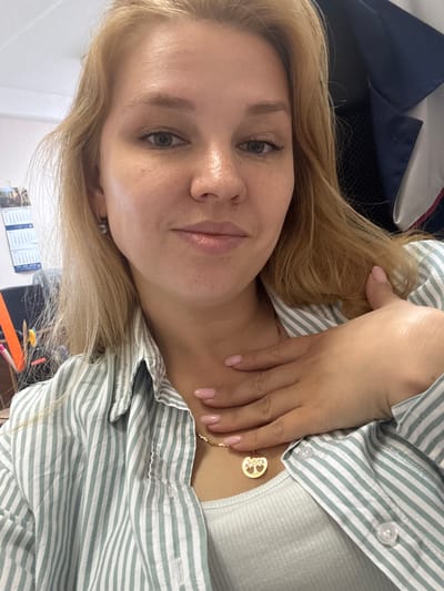 The image is a medium shot selfie of a young Caucasian woman with blonde hair, fair skin, and light green eyes. She is wearing a light green and white striped collared shirt over a white ribbed tank top. A gold necklace with a tree of life pendant is visible around her neck. Her right hand is placed on her chest, with her fingers resting near her collarbone and thumb near her neck. Her fingernails are painted a light pink color.
In the background, a blue and white jacket or fabric is visible to the right. To the left, a wall calendar with several pages is hanging, and a desk or shelf with office supplies such as pens and markers is partially visible. The environment appears to be an indoor setting, possibly an office or a room with office-like furnishings. There are no discernible landmarks or specific features that would indicate a location in the city of Заволжье, Russia. The lighting suggests an indoor environment with artificial or diffused natural light.